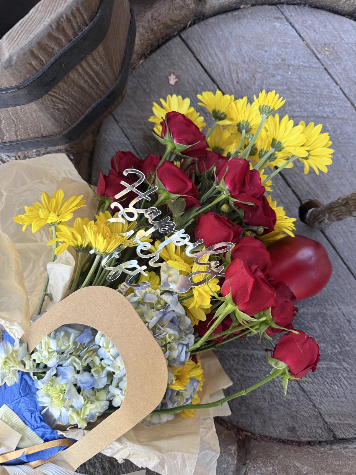 Close-up of the Fairest One bouquet with red roses, yellow daisies, pale blue hydrangea, a silver Happily Ever After pick, and a red apple accent resting on Snow White's wishing well at EPCOT's Germany pavilion