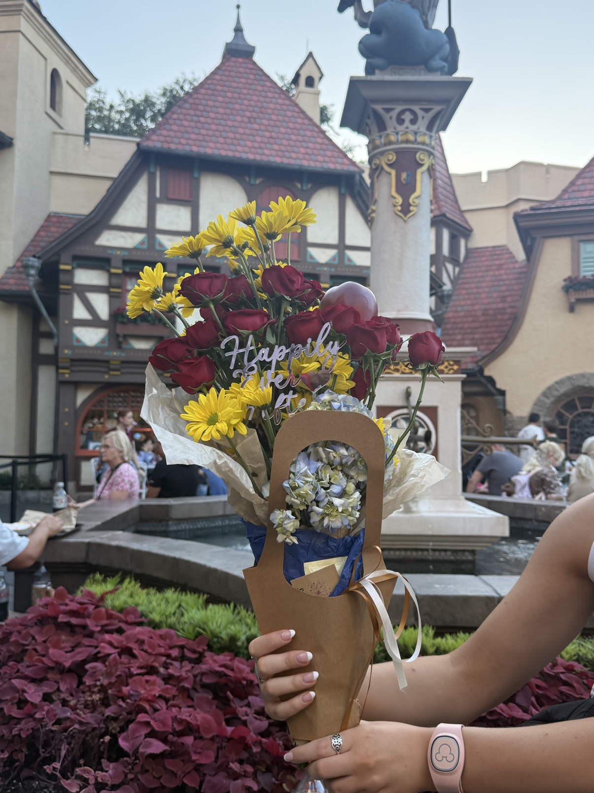 Hands holding the Fairest One bouquet by the fountain in EPCOT's Germany pavilion, with the pavilion statue behind it