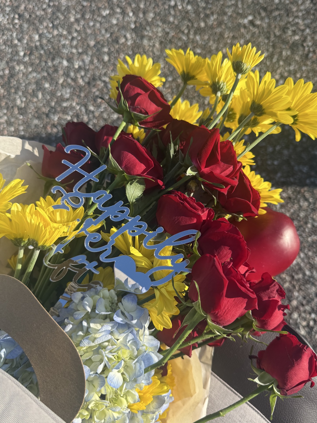 Close-up detail of red roses, yellow daisies, pale blue hydrangea, and the silver Happily Ever After pick in the Fairest One bouquet