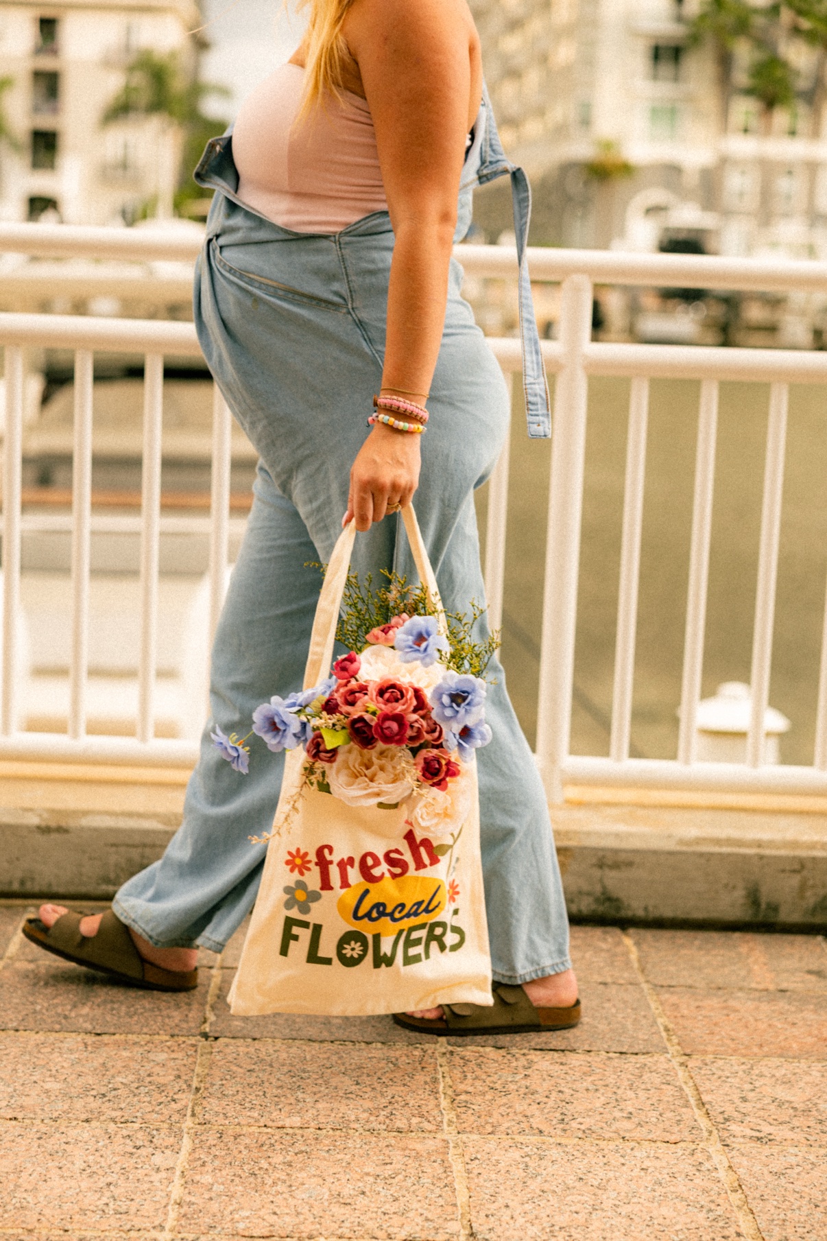 Kai walking along a waterfront railing carrying a tote bag full of fresh flowers
